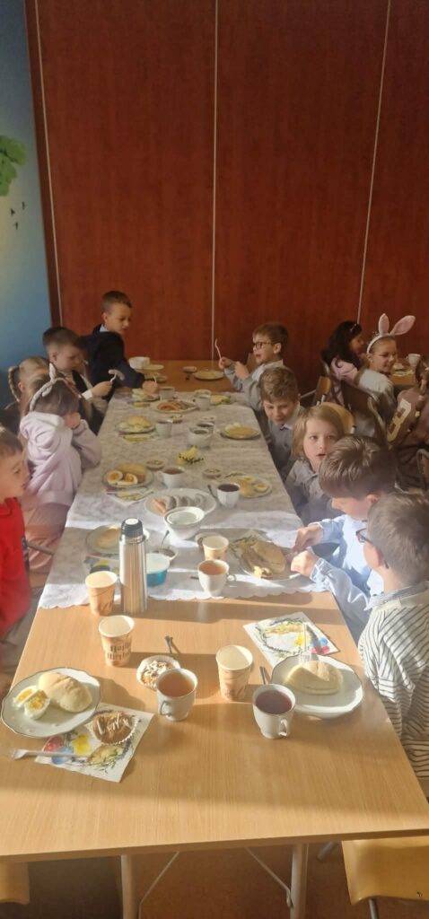 Dzieci siedzą przy długim stole podczas wspólnego posiłku; na stole znajdują się talerze z jedzeniem, kubki i termos.