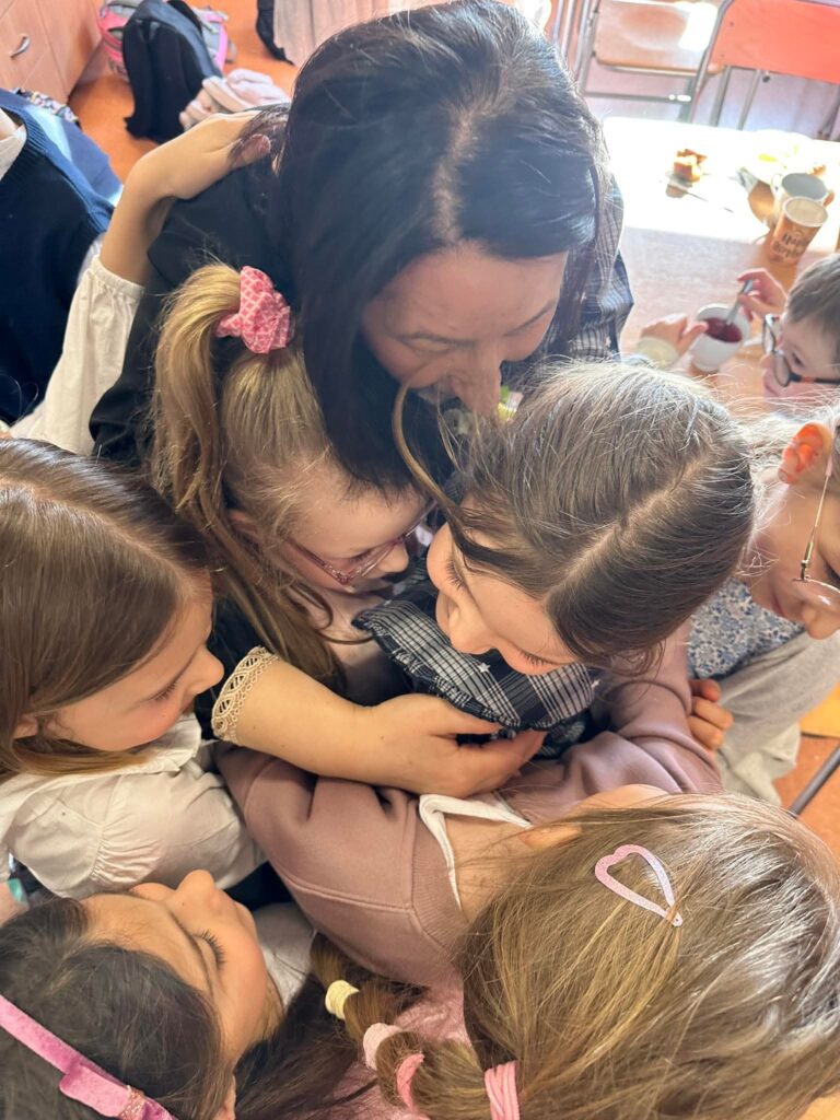 Nauczycielka przytula grupę dzieci, które otaczają ją i uśmiechają się w przyjaznej, radosnej atmosferze.