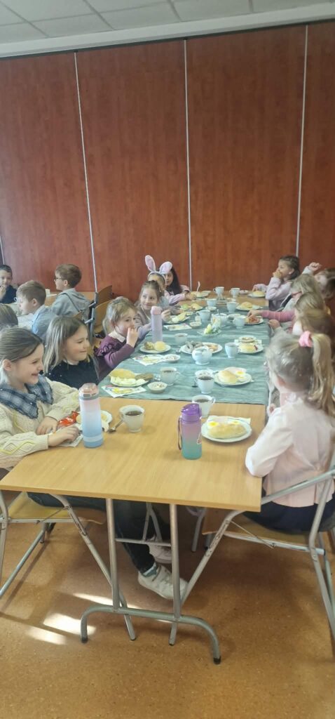 Dzieci siedzą przy długim stole i jedzą wspólne śniadanie; na stole stoją talerze z jedzeniem, kubki oraz bidony.