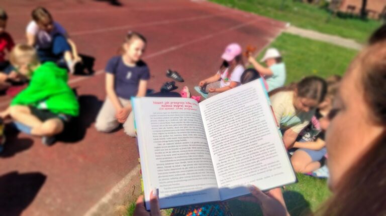Na pierwszym planie otwarta książka trzymana przez nauczyciela, w tle słuchające dzieci.