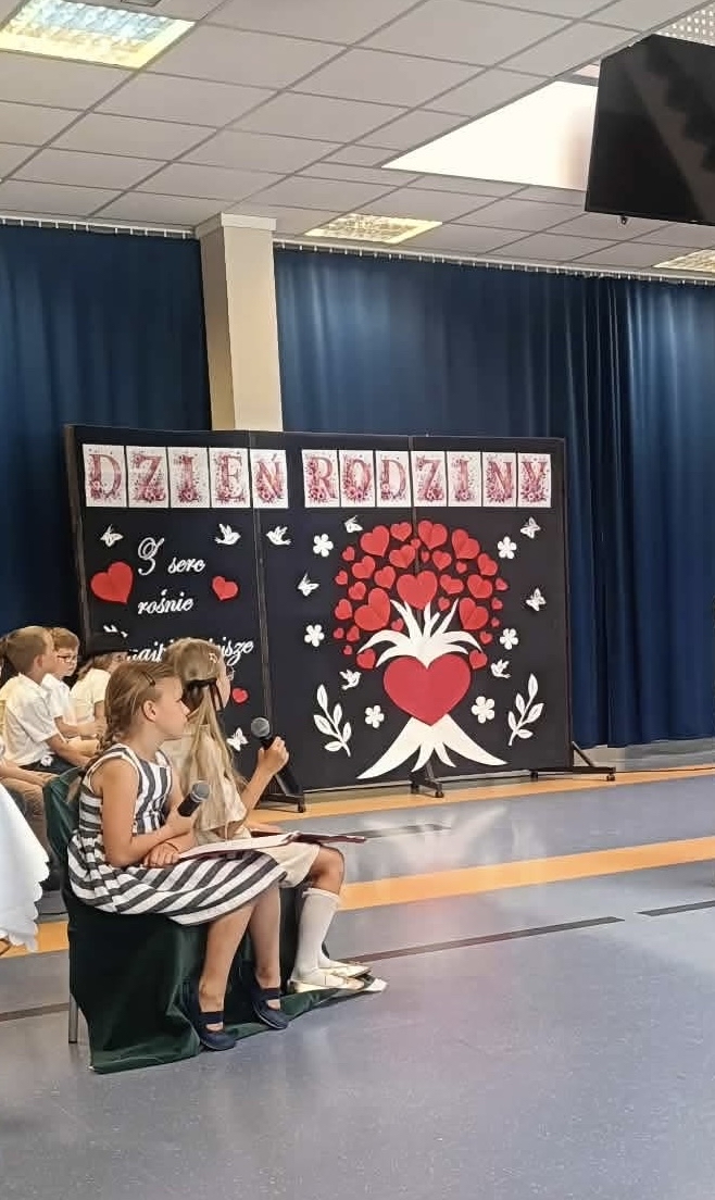 Uczniowie uczestniczą w szkolnym przedstawieniu z okazji Dnia Rodziny. Dwie dziewczynki siedzą na scenie i trzymają mikrofony, prowadząc lub prezentując część artystyczną. W tle znajduje się dekoracja z napisem „Dzień Rodziny” oraz dużym drzewem utworzonym z czerwonych serc, symbolizującym miłość, bliskość i więzi rodzinne.